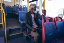 African american senior man wearing face mask sitting on bus holding smartphone. digital nomad out and about in the city during coronavirus covid 19 pandemic. — Stock Photo