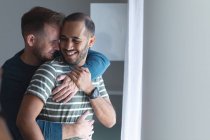 Couple masculin gay multi ethnique souriant, debout près de la fenêtre étreignant à la maison. Profiter du temps passé à la maison en isolement personnel pendant le confinement en quarantaine. — Photo de stock