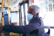 Ein älterer afroamerikanischer Mann mit Gesichtsmaske sitzt im Bus und dreht sich um, um auszusehen. digitaler Nomade unterwegs in der Stadt während Coronavirus covid 19 Pandemie. — Stockfoto