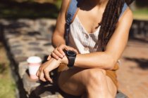 Midsection de femme afro-américaine assis en utilisant sa montre intelligente dans le parc. Numérique nomade sur le mode de vie aller. — Photo de stock