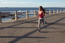 Donna afroamericana concentrata, che si allena su una passeggiata vicino al mare facendo jogging. Fitness stile di vita sano all'aperto. — Foto stock