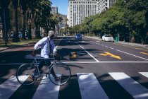 Ein afroamerikanischer Senior rast mit dem Fahrrad auf einem Fußgängerüberweg über die Straße. digitaler Nomade in der Stadt unterwegs. — Stockfoto