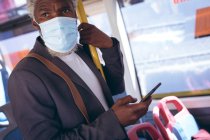 African american senior man wearing face mask standing on bus using smartphone. digital nomad out and about in the city during coronavirus covid 19 pandemic. — Stock Photo