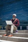 Homme âgé afro-américain assis sur des marches à l'extérieur d'un bâtiment avec du café à emporter à l'aide d'un ordinateur portable. nomade numérique dans la ville. — Photo de stock