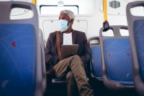 Afro-americano idoso vestindo máscara facial sentado em ônibus usando tablet digital. nômade digital para fora e sobre na cidade durante coronavírus covid 19 pandemia. — Fotografia de Stock