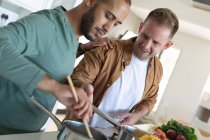 Couple masculin gay multi ethnique souriant et préparant la nourriture ensemble à la maison. Rester à la maison en isolement personnel pendant le confinement en quarantaine. — Photo de stock