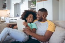 African american girl sitting on couch using digital tablet with her father. staying at home in self isolation during quarantine lockdown. — Stock Photo
