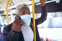 Hombre mayor afroamericano con máscara facial parado en el autobús hablando en el teléfono inteligente. nómada digital en la ciudad durante la pandemia de coronavirus covid 19. - foto de stock