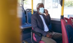 Ein älterer afroamerikanischer Mann mit Gesichtsmaske sitzt im Bus und unterhält sich per Smartphone. digitaler Nomade unterwegs in der Stadt während Coronavirus covid 19 Pandemie. — Stockfoto