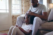 Uomo afroamericano seduto sul letto a fare i capelli alla figlia. stare a casa in isolamento durante l'isolamento in quarantena. — Foto stock
