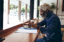 Старший африканский американец, сидящий за столом в кафе и работающий на ноутбуке. цифровая реклама в городе. — стоковое фото
