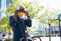 African american senior man wearing face mask sitting on bicycle in street putting on earphones. digital nomad out and about in the city during coronavirus covid 19 pandemic. — Stock Photo