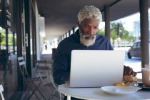 Старший африканский американец, сидящий за столом возле кафе с ноутбуком. цифровая реклама в городе. — стоковое фото