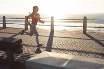 Mujer afroamericana haciendo ejercicio en un paseo junto al mar trotando. fitness estilo de vida saludable al aire libre. - foto de stock