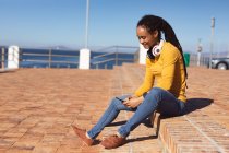 Mujer afroamericana sonriendo, usando auriculares sentados usando un teléfono inteligente en el paseo marítimo. Nómada digital sobre la marcha estilo de vida. - foto de stock