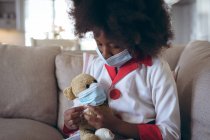 Menina afro-americana jogando médico e paciente com seu ursinho de pelúcia. ficar em casa em auto-isolamento durante o confinamento de quarentena. — Fotografia de Stock