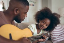 El hombre afroamericano y su hija sentados en el sofá están tocando la guitarra. permanecer en casa en aislamiento durante el bloqueo de cuarentena. - foto de stock