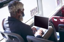 Afro-americano idoso vestindo máscara facial sentado no ônibus usando laptop. nômade digital para fora e sobre na cidade durante coronavírus covid 19 pandemia. — Fotografia de Stock