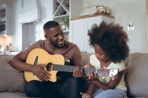 African american man and his daughter sitting on couch man is playing guitar. staying at home in self isolation during quarantine lockdown. — Stock Photo