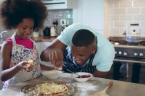 Chica afroamericana y su padre haciendo pizza juntos en la cocina. permanecer en casa en aislamiento durante el bloqueo de cuarentena. - foto de stock