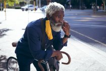 Старший африканский американец в маске, опирающийся на велосипед на улице и разговаривающий по смартфону. Цифровой кочевник в городе во время пандемии коронавируса. — стоковое фото