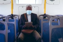 Hombre mayor afroamericano con máscara facial sentado en el autobús usando tableta digital. nómada digital en la ciudad durante la pandemia de coronavirus covid 19. - foto de stock