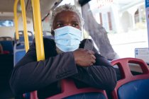 Hombre mayor afroamericano con máscara facial sentado en el autobús sosteniendo el teléfono inteligente. nómada digital en la ciudad durante la pandemia de coronavirus covid 19. - foto de stock