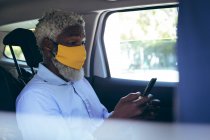 Homem sênior afro-americano usando máscara facial sentado em táxi táxi usando smartphone. nômade digital para fora e sobre na cidade durante coronavírus covid 19 pandemia. — Fotografia de Stock
