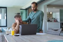 Worried multi ethnic gay male couple going through bills and using laptop at home. Staying at home in self isolation during quarantine lockdown. — Stock Photo