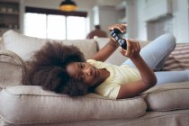 Afro-americano deitado num sofá a jogar videojogos. ficar em casa em auto-isolamento durante o confinamento de quarentena. — Fotografia de Stock
