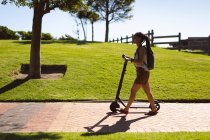 Felice donna afroamericana con lo zaino che cammina con uno scooter nel parco. Nomade digitale in movimento lifestyle. — Foto stock