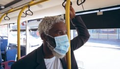Un aîné afro-américain portant un masque sur le bus regardant par la fenêtre. numérique nomade dehors et environ dans la ville pendant coronavirus covid 19 pandémie. — Photo de stock