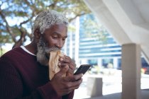 Африканський старший американець на вулиці їсть сендвіч і користується смартфоном. Цифровий кочівник і все в місті. — стокове фото