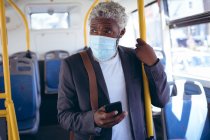 Homme âgé afro-américain portant un masque facial debout sur un bus tenant un smartphone. numérique nomade dehors et environ dans la ville pendant coronavirus covid 19 pandémie. — Photo de stock