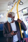 Homme âgé afro-américain portant un masque facial debout sur un bus tenant un smartphone. numérique nomade dehors et environ dans la ville pendant coronavirus covid 19 pandémie. — Photo de stock