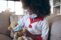 Menina afro-americana jogando médico e paciente com seu ursinho de pelúcia. ficar em casa em auto-isolamento durante o confinamento de quarentena. — Fotografia de Stock