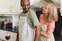 Разнообразная старшая пара надевает фартуки на кухне перед готовкой. оставаться дома в изоляции во время карантинной изоляции. — стоковое фото