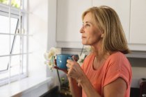 Caucasian senior woman standing by window drinking cup of coffee. staying at home in isolation during quarantine lockdown. — Stock Photo