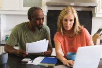 Diverse senior couple sitting in kitchen using laptop and going through paperwork. staying at home in isolation during quarantine lockdown. — Stock Photo
