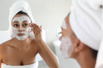 Afroamerikanerin trägt Gesichtsmaske auf, während sie im Badezimmer in den Spiegel schaut. Zu Hause bleiben in Selbstisolierung in Quarantäne — Stockfoto
