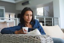 Mulher de raça mista relaxando na sala de estar no sofá com uma xícara de café. ficar em casa em isolamento durante o confinamento de quarentena. — Fotografia de Stock