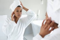 Mujer afroamericana en albornoz tocándose la cara mientras se mira en el espejo en el baño. permaneciendo en casa en aislamiento en cuarentena - foto de stock