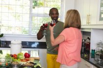 Diverse senior couple standing in kitchen making a toast with glasses of wine. staying at home in isolation during quarantine lockdown. — Stock Photo