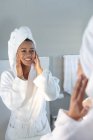 Mujer afroamericana sonriente en albornoz tocándose la cara mientras se mira en el espejo en el baño. permaneciendo en casa en aislamiento en cuarentena - foto de stock