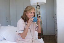 Mujer mayor caucásica sentada en la cama y tomando una taza de café. permanecer en casa aislado durante el bloqueo de cuarentena. - foto de stock