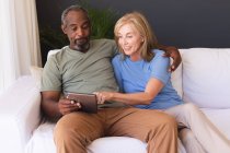 Разнообразная пожилая пара, сидящая на диване и пользующаяся цифровым планшетом. оставаться дома в изоляции во время карантинной изоляции. — стоковое фото