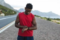 Un homme afro-américain faisant de l'exercice en plein air vérifiant sa montre connectée sur une route côtière. entraînement physique et mode de vie sain en plein air. — Photo de stock