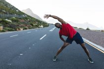 Homem afro-americano exercitando ao ar livre que se estende em uma estrada costeira. treinamento de fitness e estilo de vida saudável ao ar livre. — Fotografia de Stock