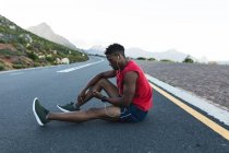 Homme afro-américain faisant de l'exercice à l'extérieur en utilisant un smartphone sur une route côtière. entraînement physique et mode de vie sain en plein air. — Photo de stock