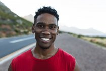 Porträt eines fitten afrikanisch-amerikanischen Mannes, der an einer Küstenstraße im Freien trainiert und in die Kamera lächelt. Fitnesstraining und gesunder Lebensstil im Freien. — Stockfoto